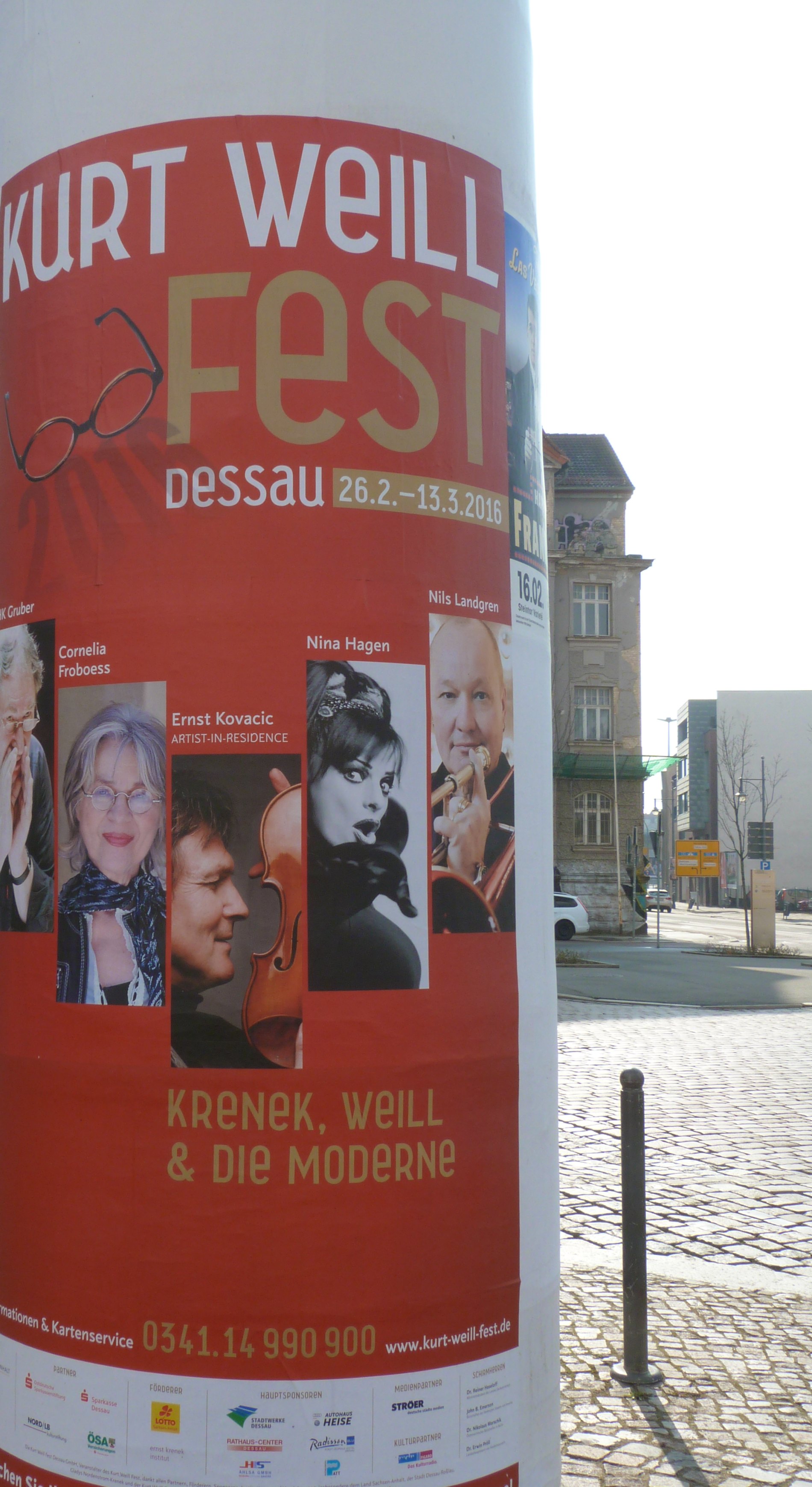Advertising column with a large red Kurt Weill Fest poster listing performers and dates, cobbled square behind.